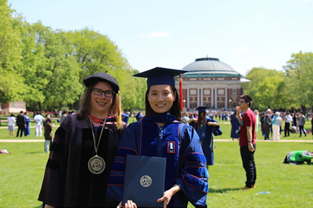 Cathy Murphy and Jingyu Huang in their caps and gowns on the quad with the Foellinger Auditorium in the background for Jingyu's graduation, May 2014.